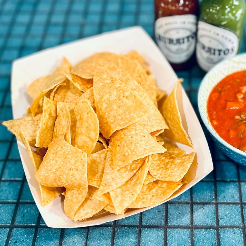 A white tray of tortilla chips, a bowl of salsa, and two hot sauce bottles on a blue tiled surface.
