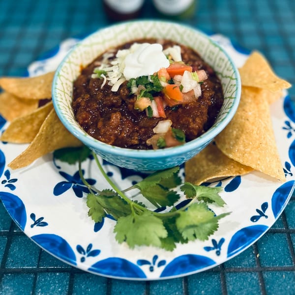 A bowl of chili with sour cream, pico de gallo, and shredded cheese, surrounded by tortilla chips.