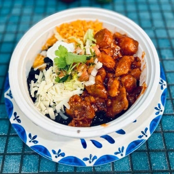 A white bowl filled with orange chicken, rice, black beans, shredded cheese, pico de gallo, and sour cream.